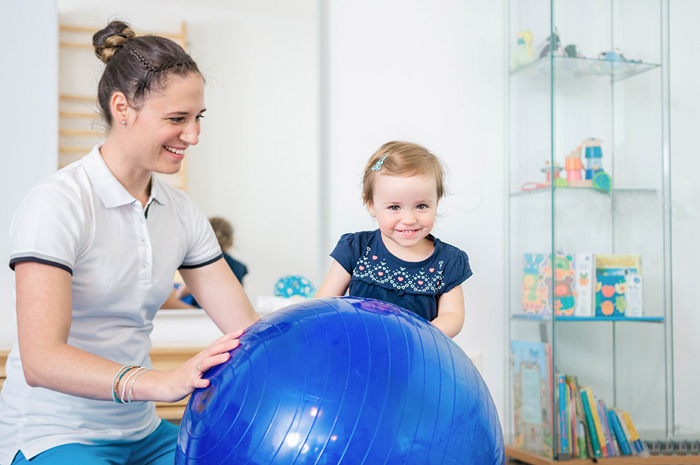Kind hinter einem großen blauen Ball und Julia Födinger als Therapeut